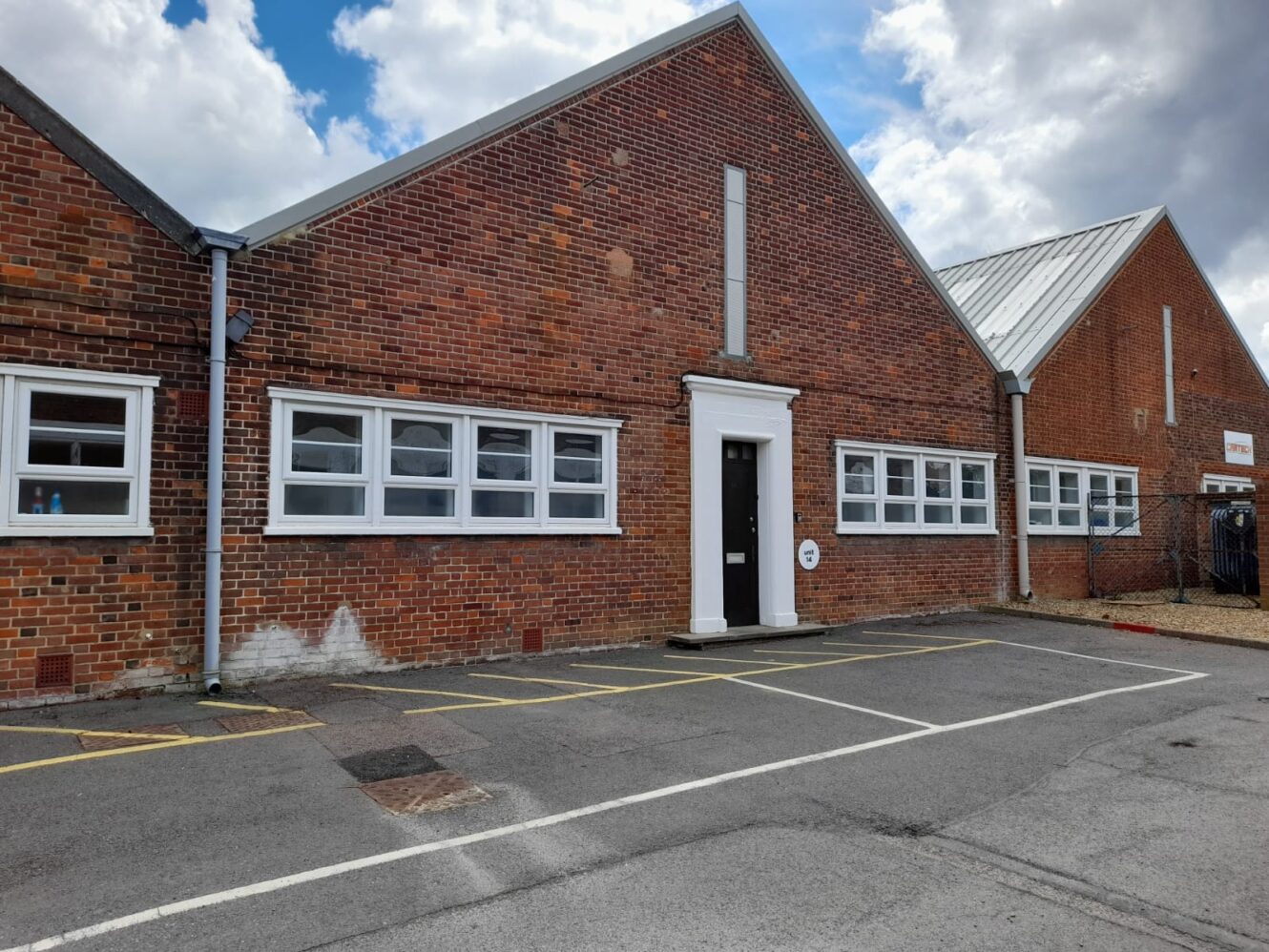 Brick building with multiple white-framed windows and a central black door, set against a cloudy sky, with a parking area in the foreground.