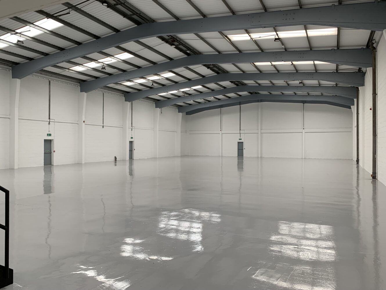 Empty industrial warehouse with white walls, gray steel beams, and a polished concrete floor, illuminated by natural light from windows in the ceiling.