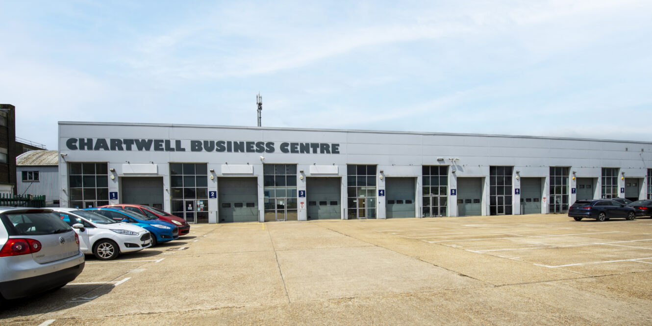A row of parked cars stands in front of the Chartwell Business Centre, a large, gray industrial building with multiple garage-style doors and clear signage.