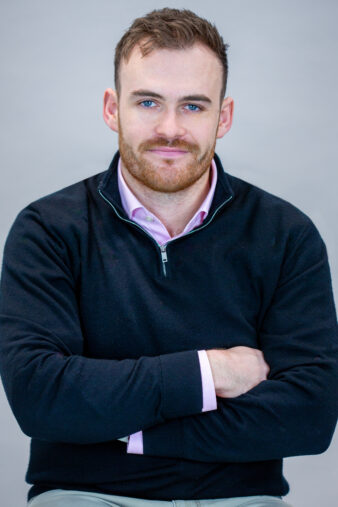 A man with short brown hair and a beard, wearing a black quarter-zip sweater over a pink collared shirt, sits with arms crossed against a plain background.
