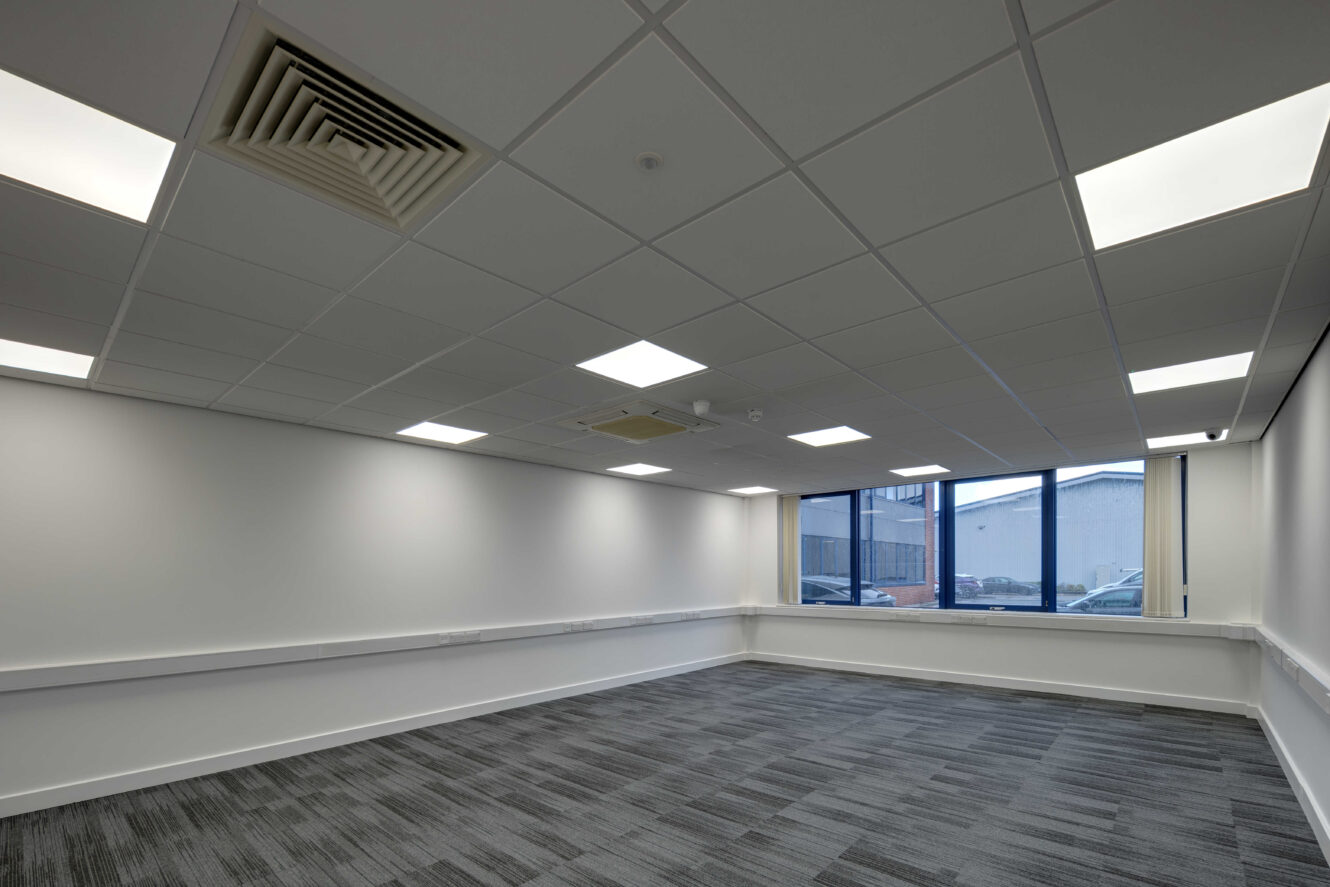 A modern, empty conference room with grey carpet, white walls, a drop ceiling with fluorescent lights, and large windows facing an industrial building.