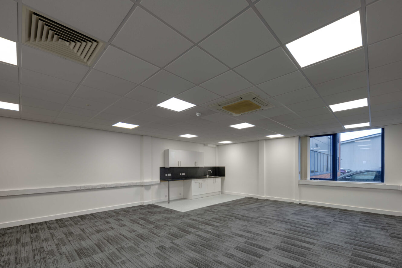 Unfurnished office space with gray carpet, white walls, ceiling lights, a kitchenette area, and a large window.
