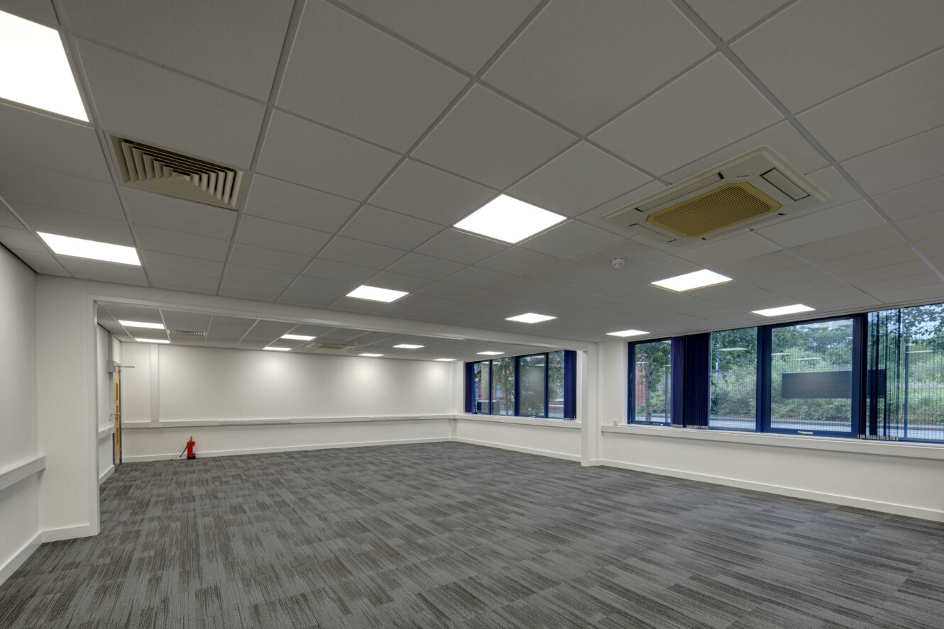 An empty office room with gray carpet, white walls, large windows on one side, and a tiled ceiling with fluorescent lighting.