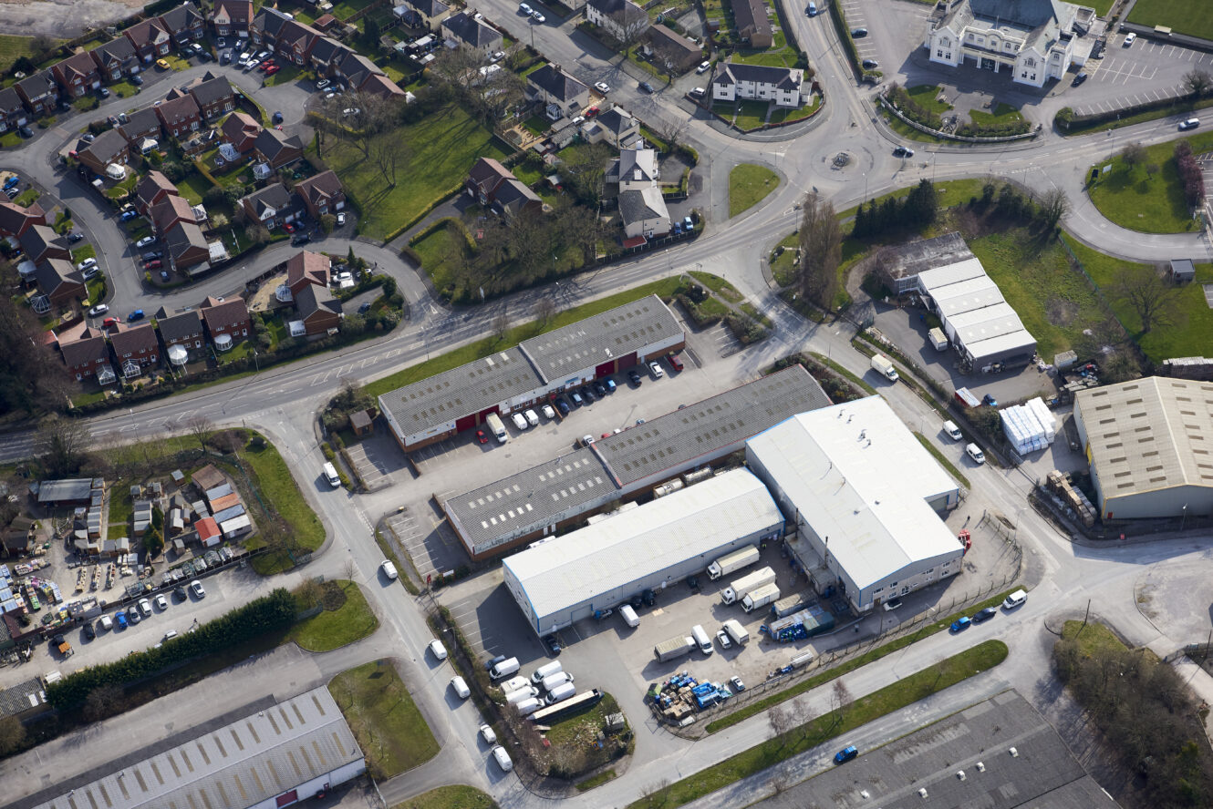 Aerial view of an industrial area with warehouses, trucks, parked vehicles, nearby roads, and adjacent residential neighborhood.