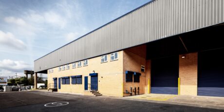 A large industrial warehouse with yellow brick walls, blue roller doors, small windows, and marked parking spaces outside under a partly cloudy sky.