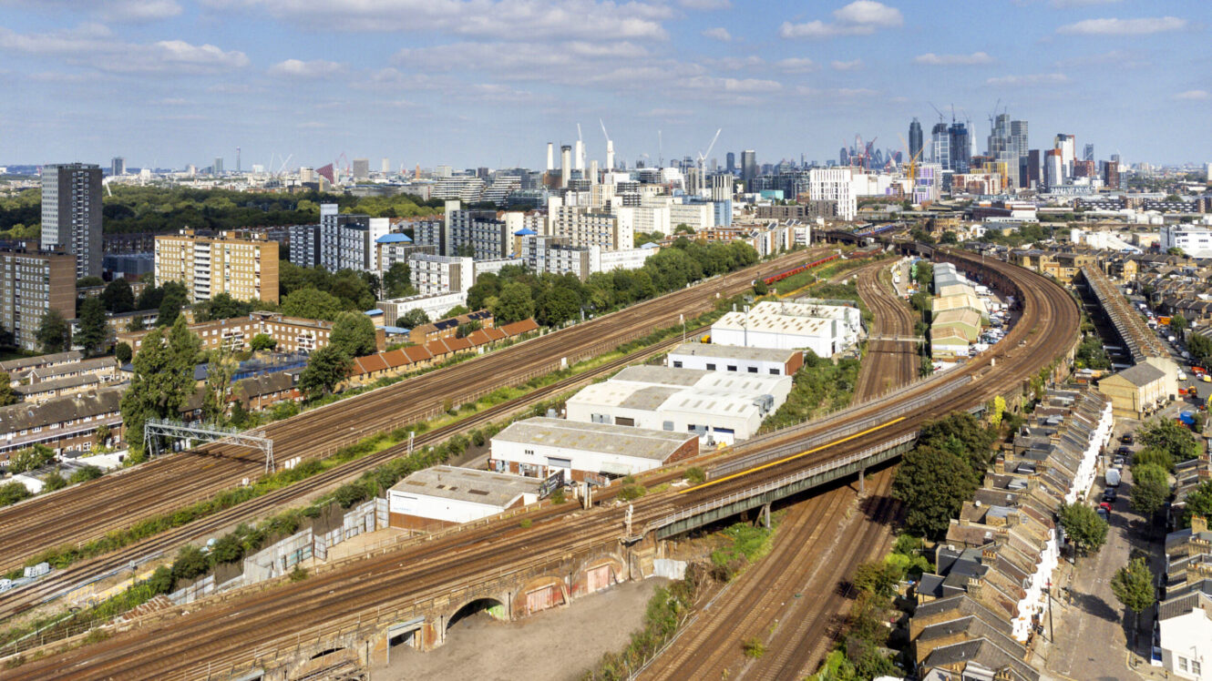 Aerial view of multiple railway tracks running through an urban area with residential and commercial buildings; city skyline visible in the distance under a partly cloudy sky.