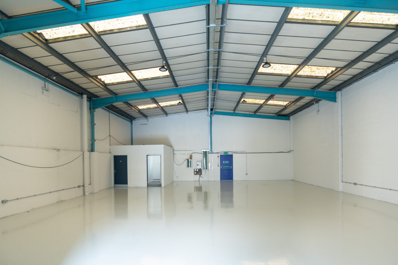 Interior of an empty warehouse with white walls, polished floor, blue steel beams, and a small partitioned room with two doors. Overhead lights and skylights are visible.
