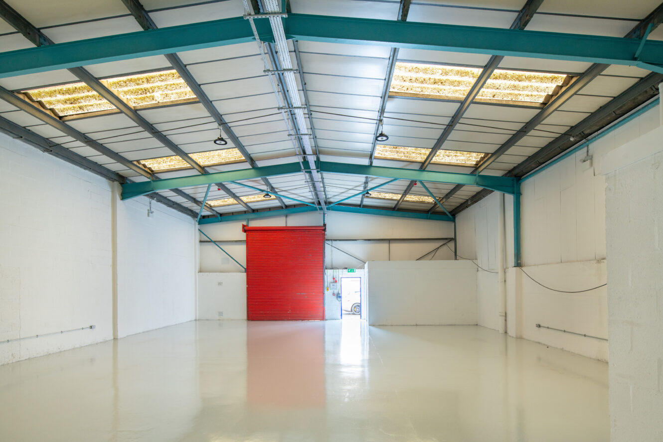 Empty industrial warehouse interior with white walls, polished floor, exposed steel beams, and a partially open red roller shutter door letting in natural light.