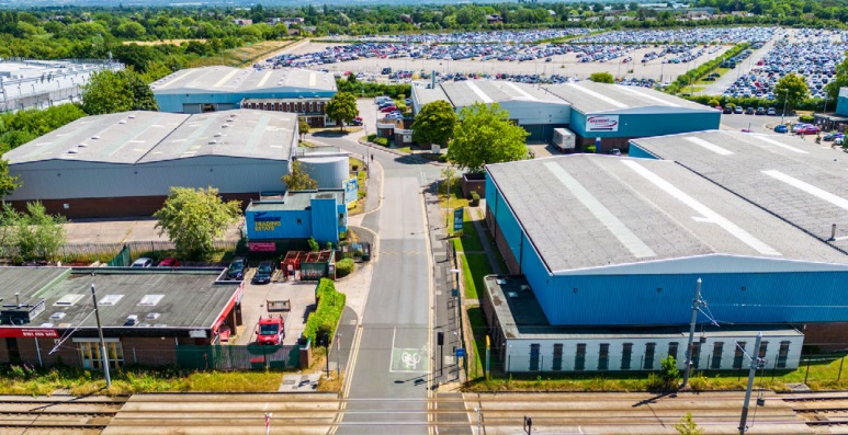 Aerial view of an industrial estate with large warehouses, a few parked vehicles, and a nearby parking lot filled with cars, bordered by greenery in the background.