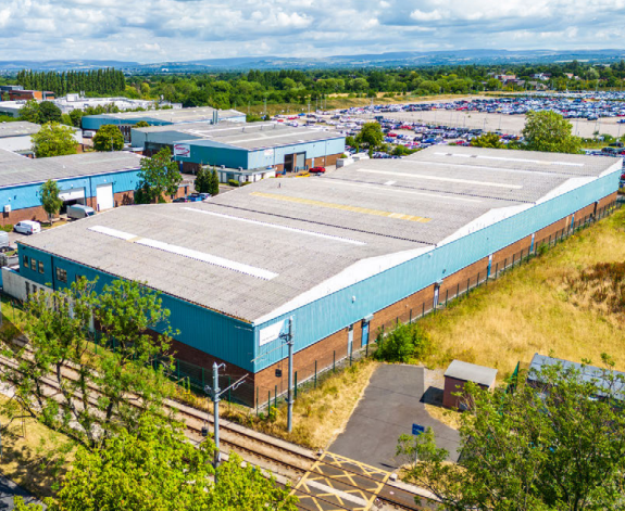 A large industrial warehouse with a blue and brick exterior, surrounded by fencing, near railway tracks and other industrial buildings, with a parking lot and green landscape in the distance.