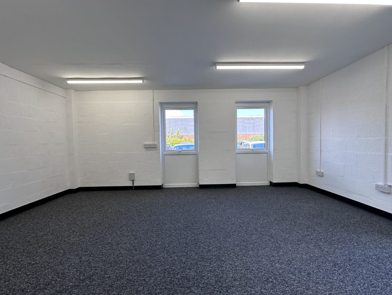 Empty office room with white walls, two windows, grey carpeted floor, and ceiling lights. Cars and a building are visible through the windows.