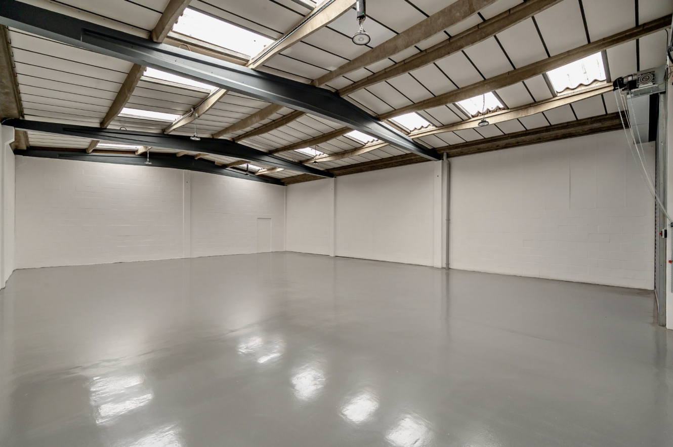 Empty industrial warehouse space with white walls, high ceiling, exposed beams, and polished concrete floor, illuminated by skylights and overhead lights.