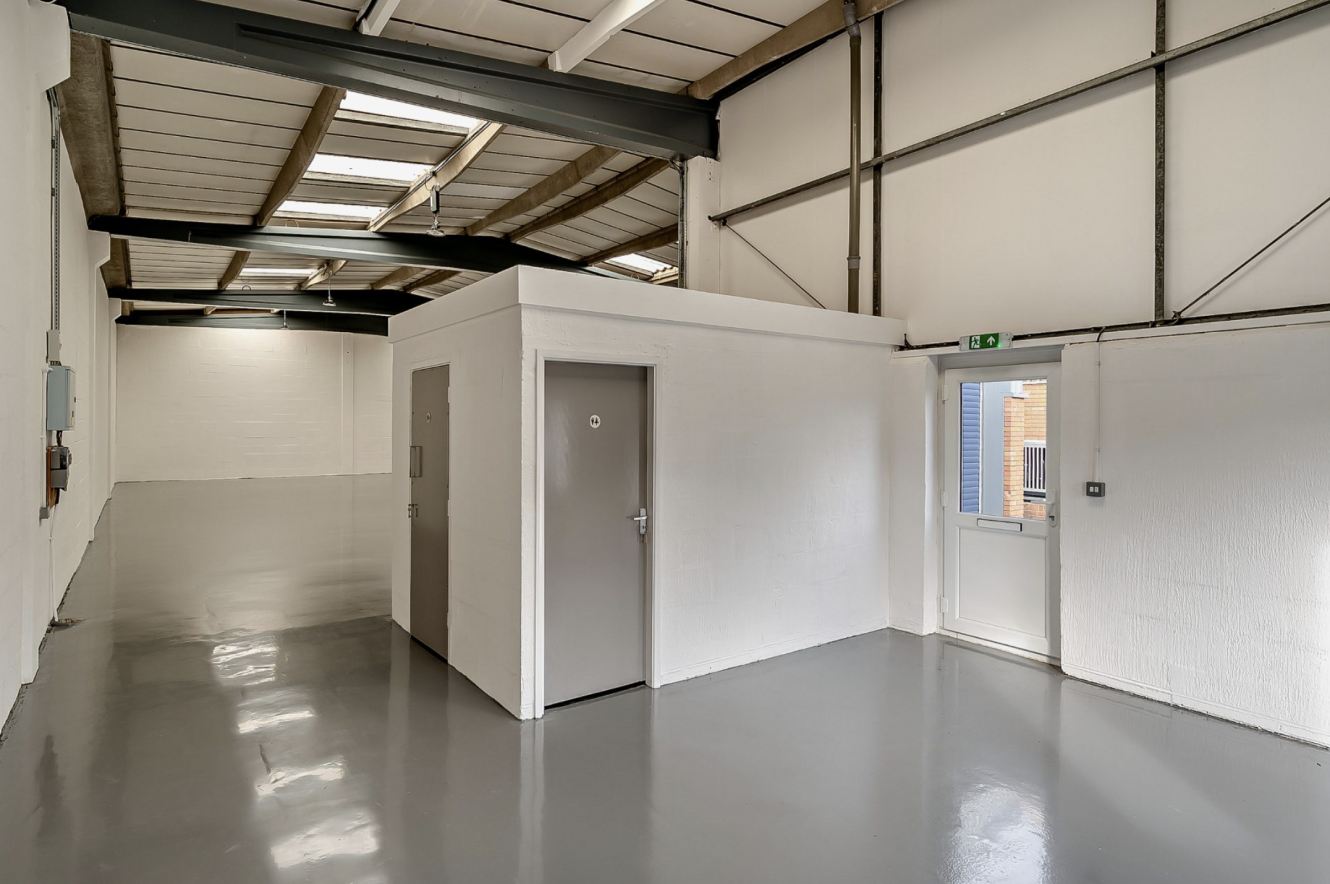 Interior of an empty industrial warehouse with white walls, gray doors, polished concrete floor, and a small enclosed room near the entrance. Natural light comes from windows in the ceiling.