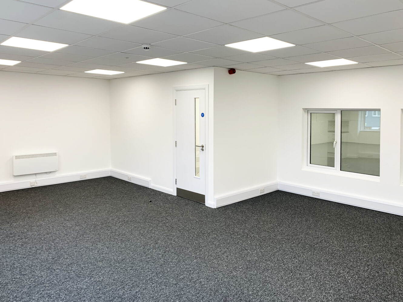 Empty office room with white walls, gray carpet, a closed door, a wall heater, a small window, and a ceiling with rectangular fluorescent lights.