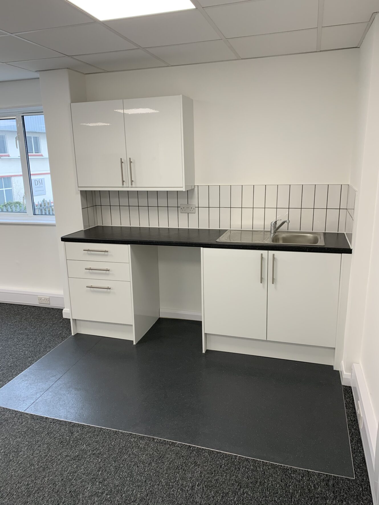 Small modern kitchenette with white cabinets, black countertop, stainless steel sink, tiled backsplash, and a window to the left. The floor changes from carpet to vinyl near the kitchenette.