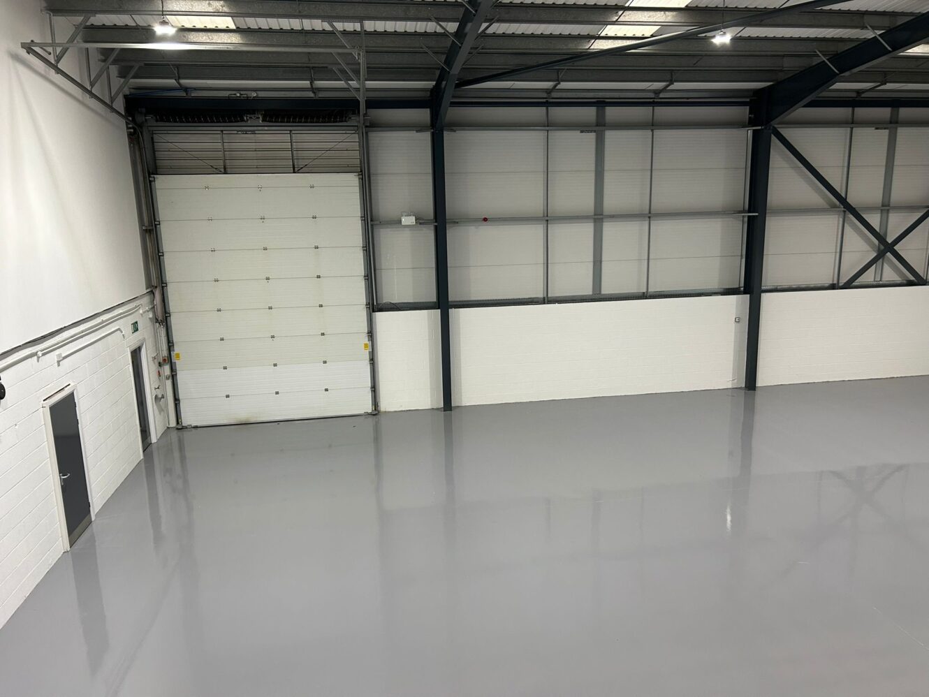 Interior of an empty industrial warehouse with a white roller door, high ceiling, and smooth, shiny grey floor.