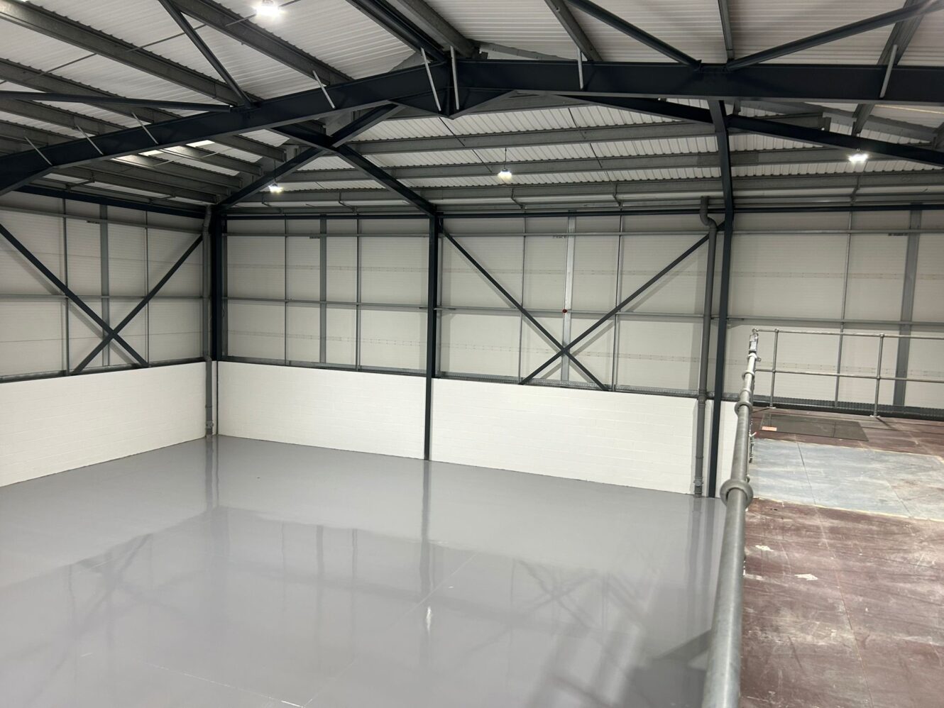 Large, empty industrial warehouse space with grey epoxy flooring, white lower walls, exposed metal beams, and bright overhead lighting.