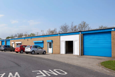 A row of industrial storage units with blue doors, several parked cars, and an open unit door under a clear sky.