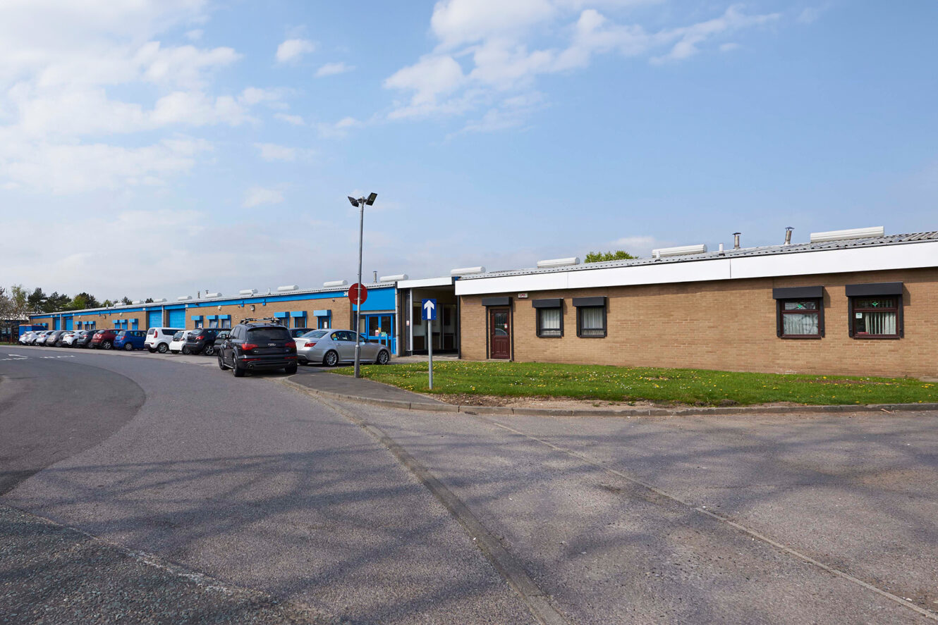 Single-story office and industrial building with parked cars along the street under a partly cloudy sky.