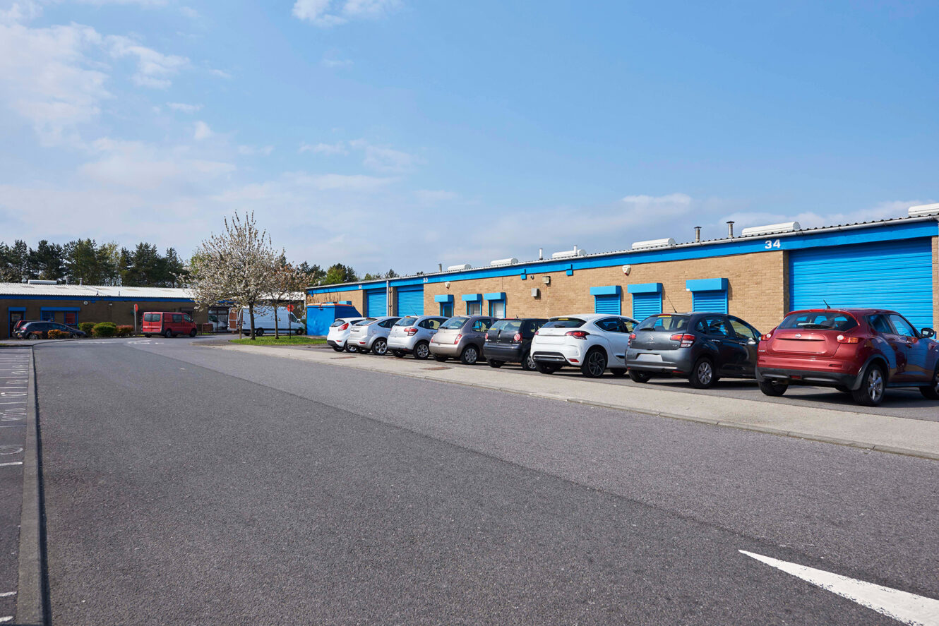 Row of cars parked in front of a low industrial building with blue doors and shutters on a clear day.