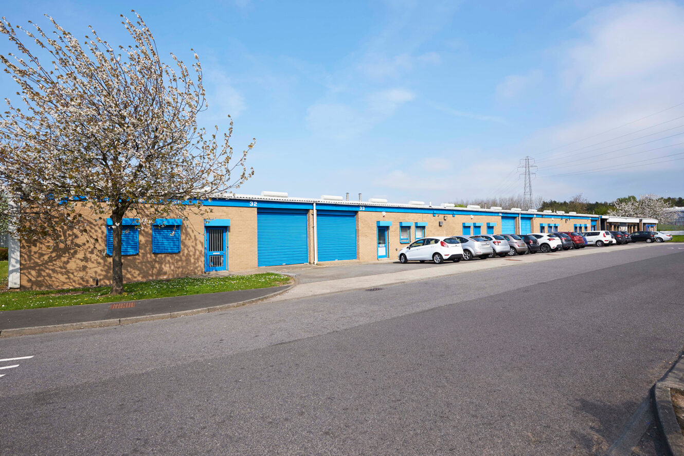 Single-story industrial building with blue doors and windows, several parked cars in front, and a flowering tree on the left under a partly cloudy sky.
