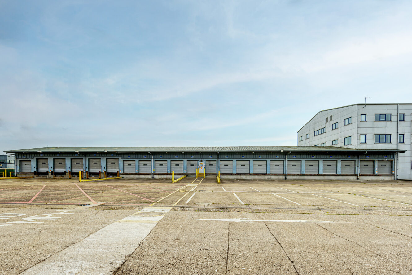 A large warehouse building with multiple loading docks and an adjacent office section, viewed from an empty parking lot under a clear sky.