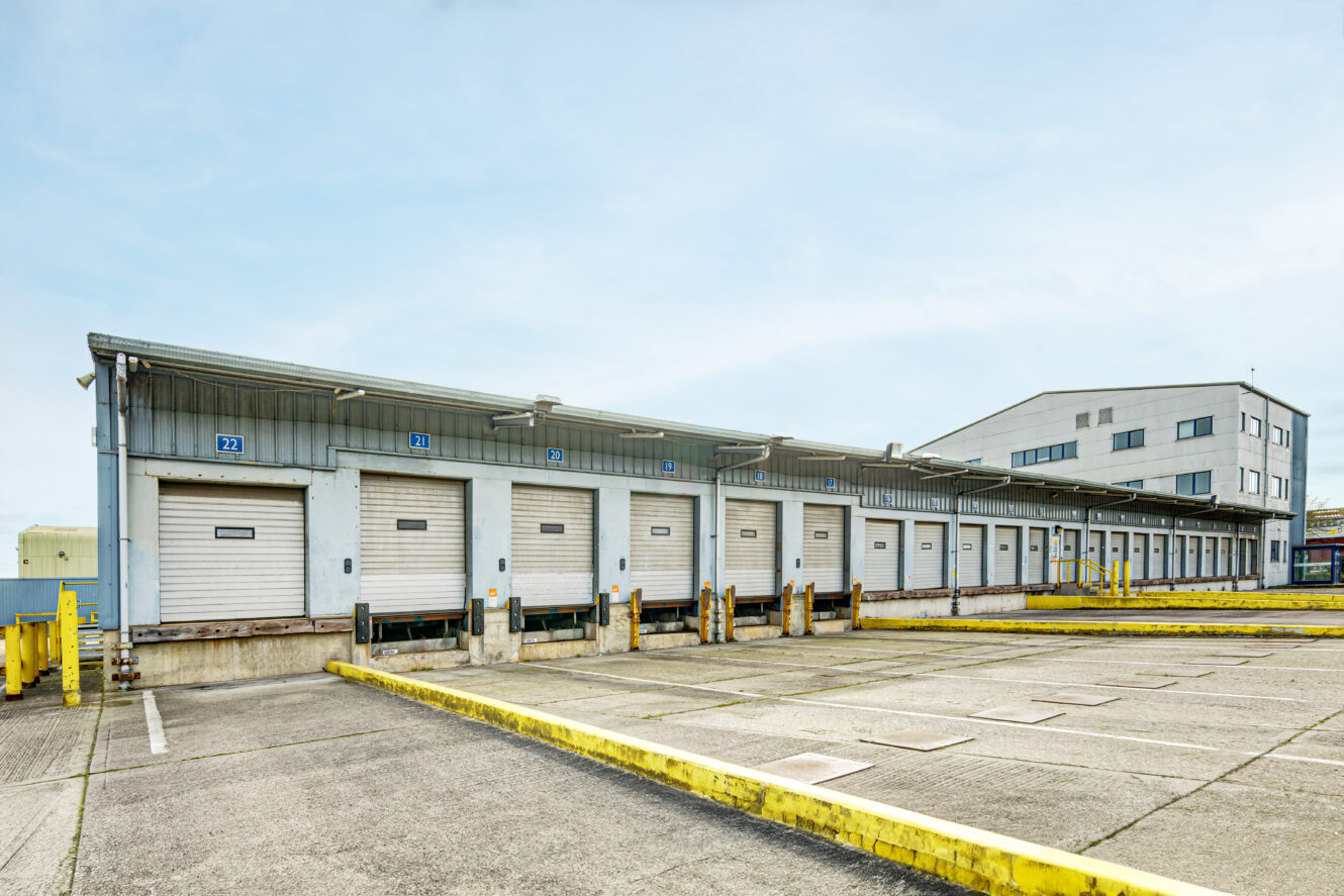 A row of closed loading dock doors at an industrial warehouse with yellow safety barriers and empty parking spaces in front.