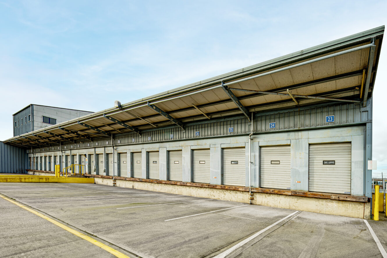 A row of closed loading dock doors with overhead covering at an industrial warehouse, with empty parking spaces in front.
