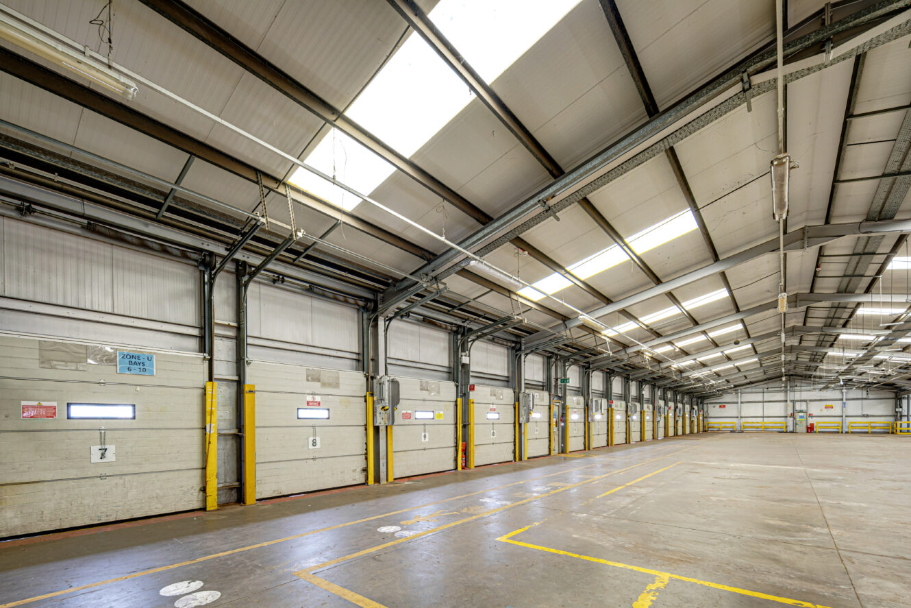 Interior of an empty warehouse with high ceilings, large loading dock doors, overhead lighting, and marked parking spaces on the concrete floor.