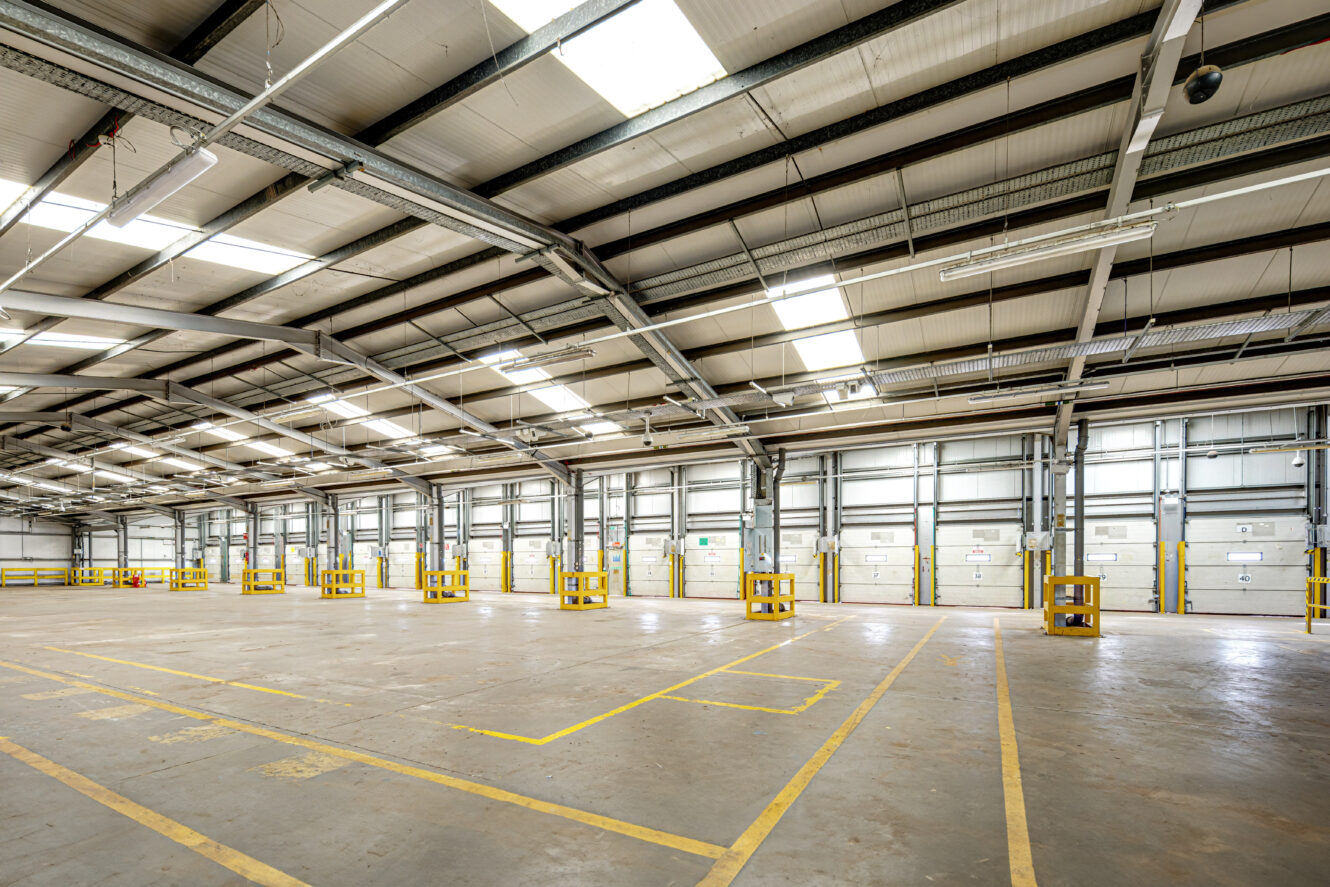 Wide, empty industrial warehouse interior with high ceilings, exposed beams, skylights, and marked parking bays on the concrete floor.