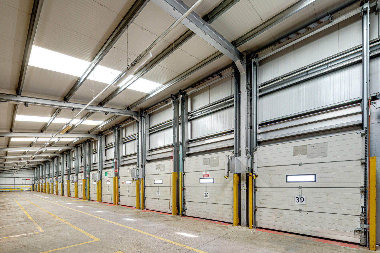 Interior of an empty industrial loading dock with multiple closed loading bay doors, numbered 38 and 39, yellow safety barriers, and overhead lighting.