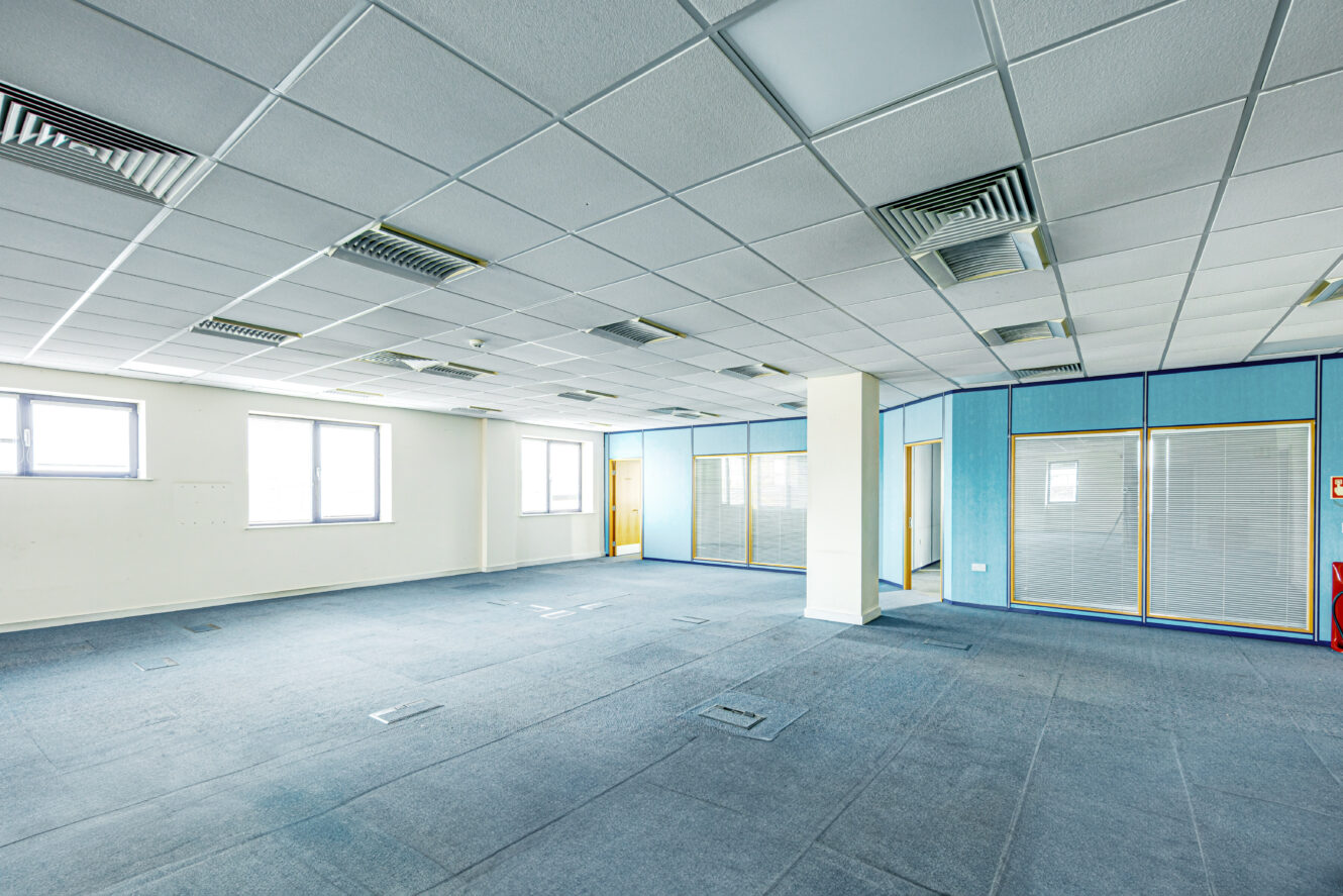 Empty office space with carpeted floor, large windows, suspended ceiling tiles, blue partition walls, and fluorescent lighting.