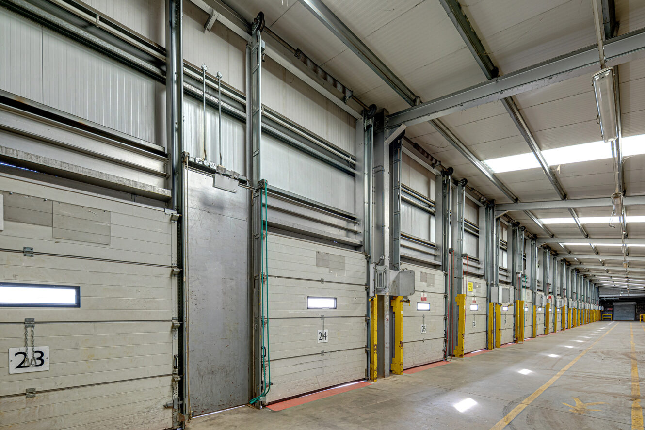 Interior view of an empty warehouse loading dock with multiple closed bay doors, concrete floor, and numbered sections on each door.