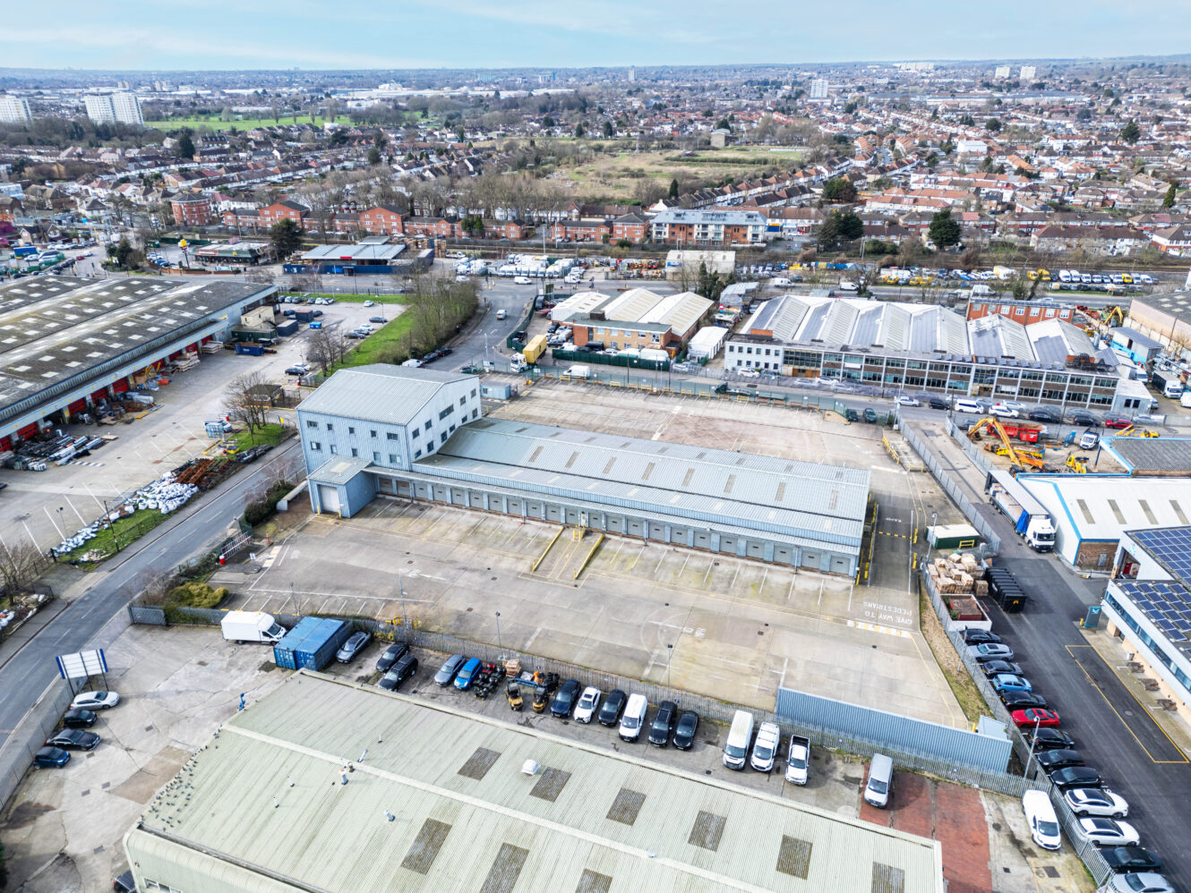 Aerial view of an industrial area with warehouses, parked cars, and surrounding residential neighborhoods under a partly cloudy sky.