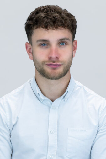 A young man with short curly hair and a beard, wearing a white button-up shirt, poses against a plain light background.