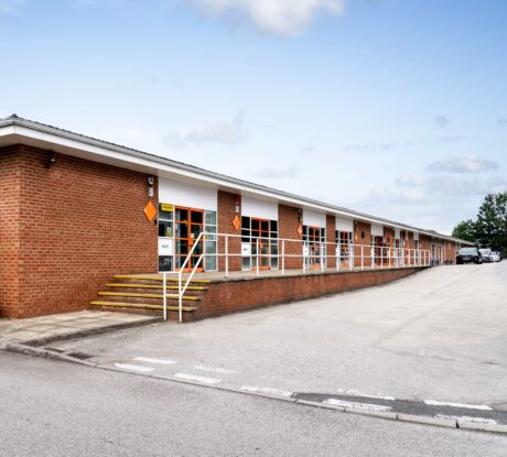 A row of single-story brick commercial units with orange doors and windows, white railings, and an empty parking lot in front.
