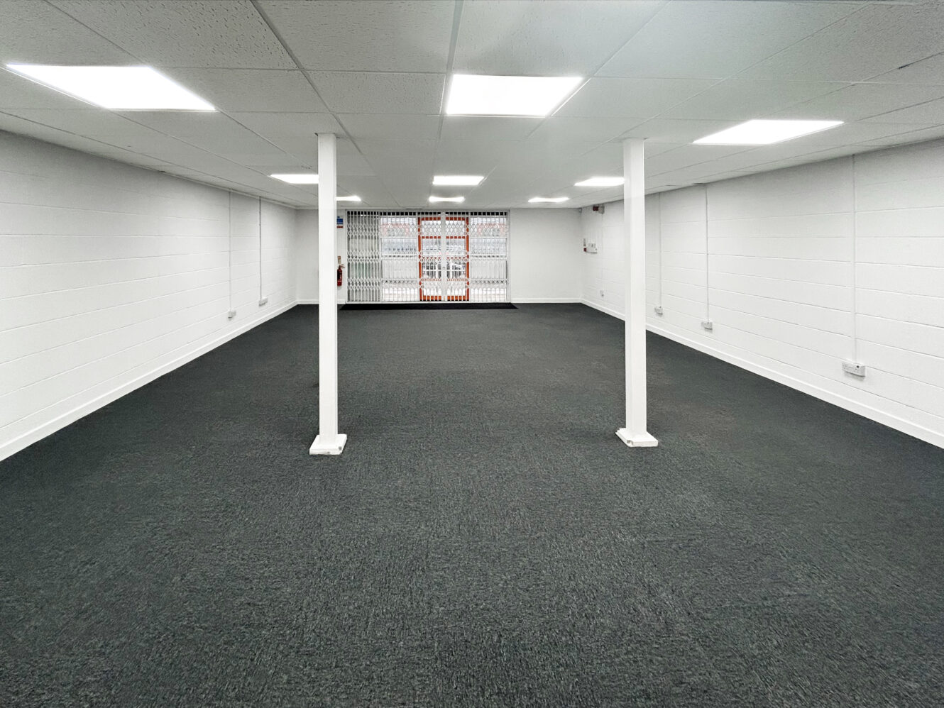 Empty room with gray carpet, white walls, ceiling lights, support columns, and a grated window at the far end.