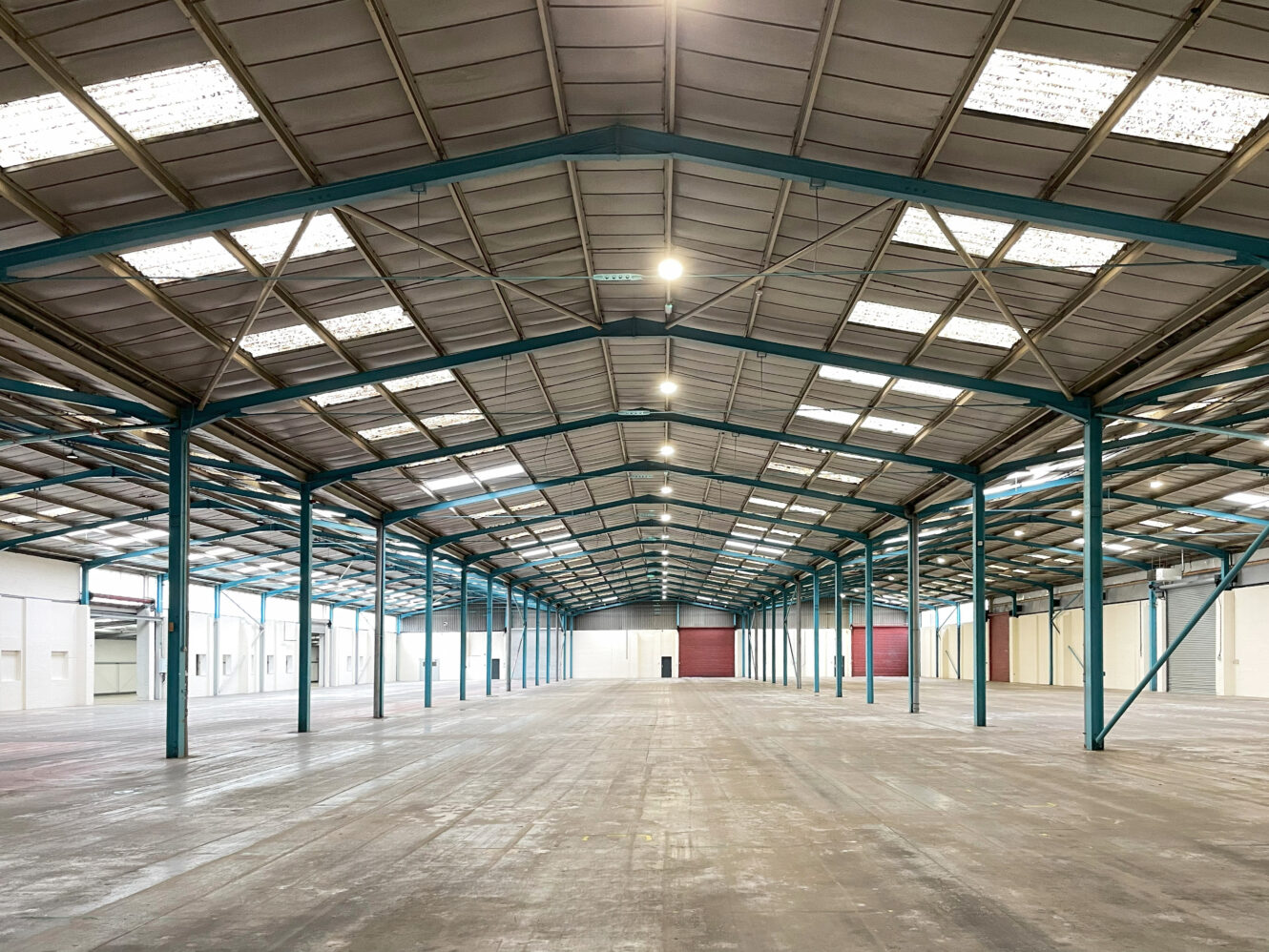 Interior of a large, empty warehouse with a high ceiling, exposed steel beams, and rows of skylights. The floor is concrete, and the space is well-lit and spacious.