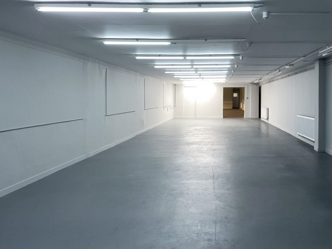 Empty room with plain white walls, grey floors, and fluorescent ceiling lights, with a doorway visible at the far end.