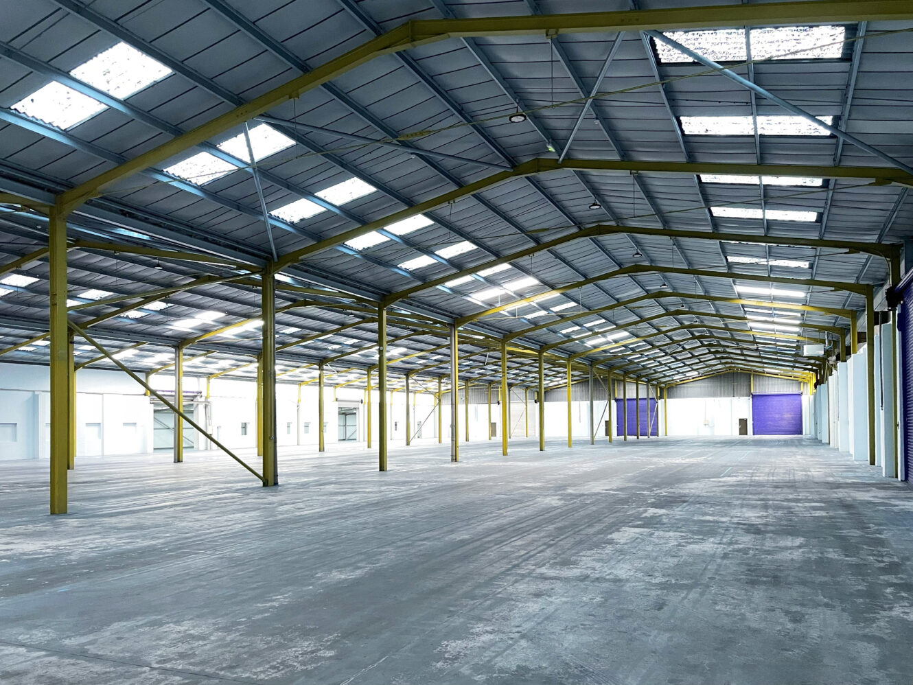 Interior of a large, empty warehouse with a high ceiling, metal beams, skylights, concrete floor, and wide entry doors.