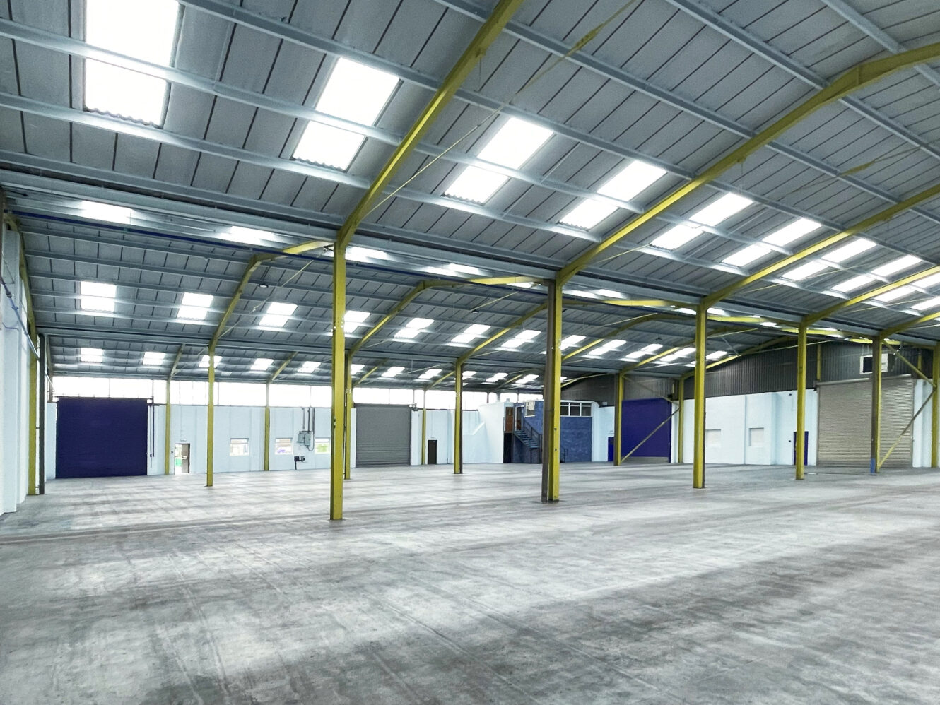 Large empty warehouse interior with high ceilings, yellow support beams, overhead lighting, and concrete floor.