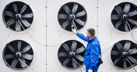 A technician in a blue uniform inspects large industrial fans mounted on a wall, using a handheld measuring device.