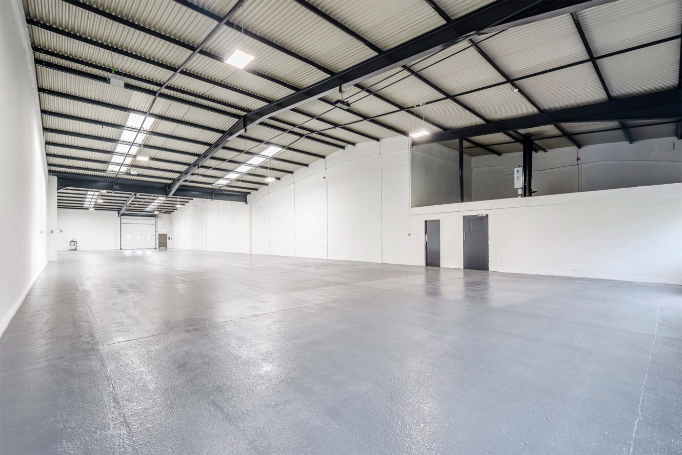 Large, empty industrial warehouse with polished gray concrete floor, white walls, high ceiling, exposed beams, and doors along the right wall.