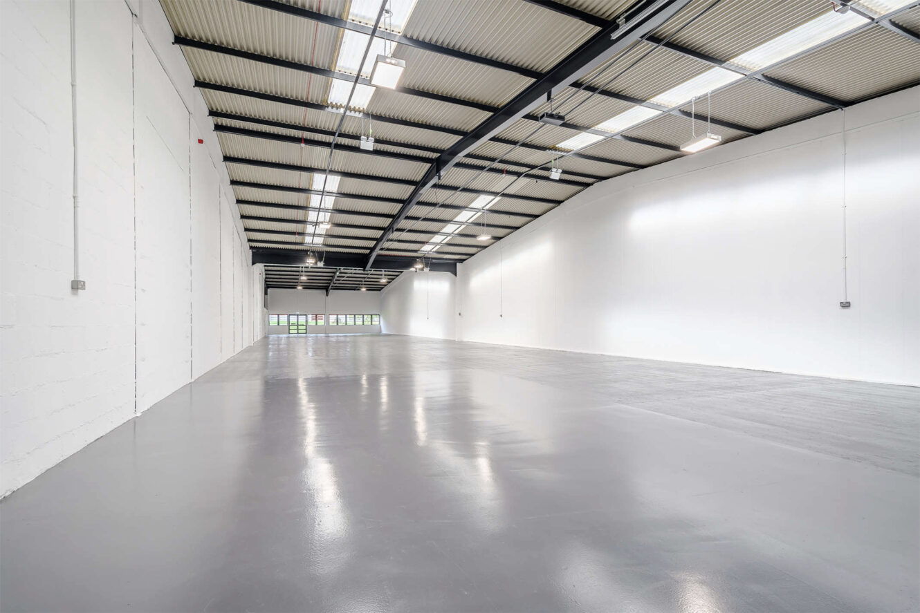 Large empty warehouse with high ceilings, white walls, exposed beams, polished gray concrete floor, and industrial lighting.