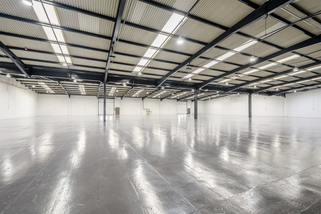 Large empty warehouse with polished concrete floor, high ceiling, exposed beams, and bright overhead lighting. White walls and wide open space dominate the scene.