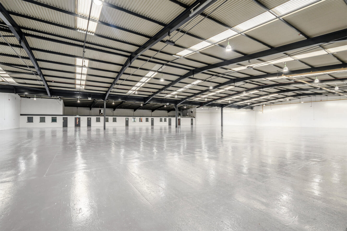 Large, empty industrial warehouse with polished concrete floor, high ceiling, exposed beams, and rows of fluorescent lights. White walls and small windows line the far side.