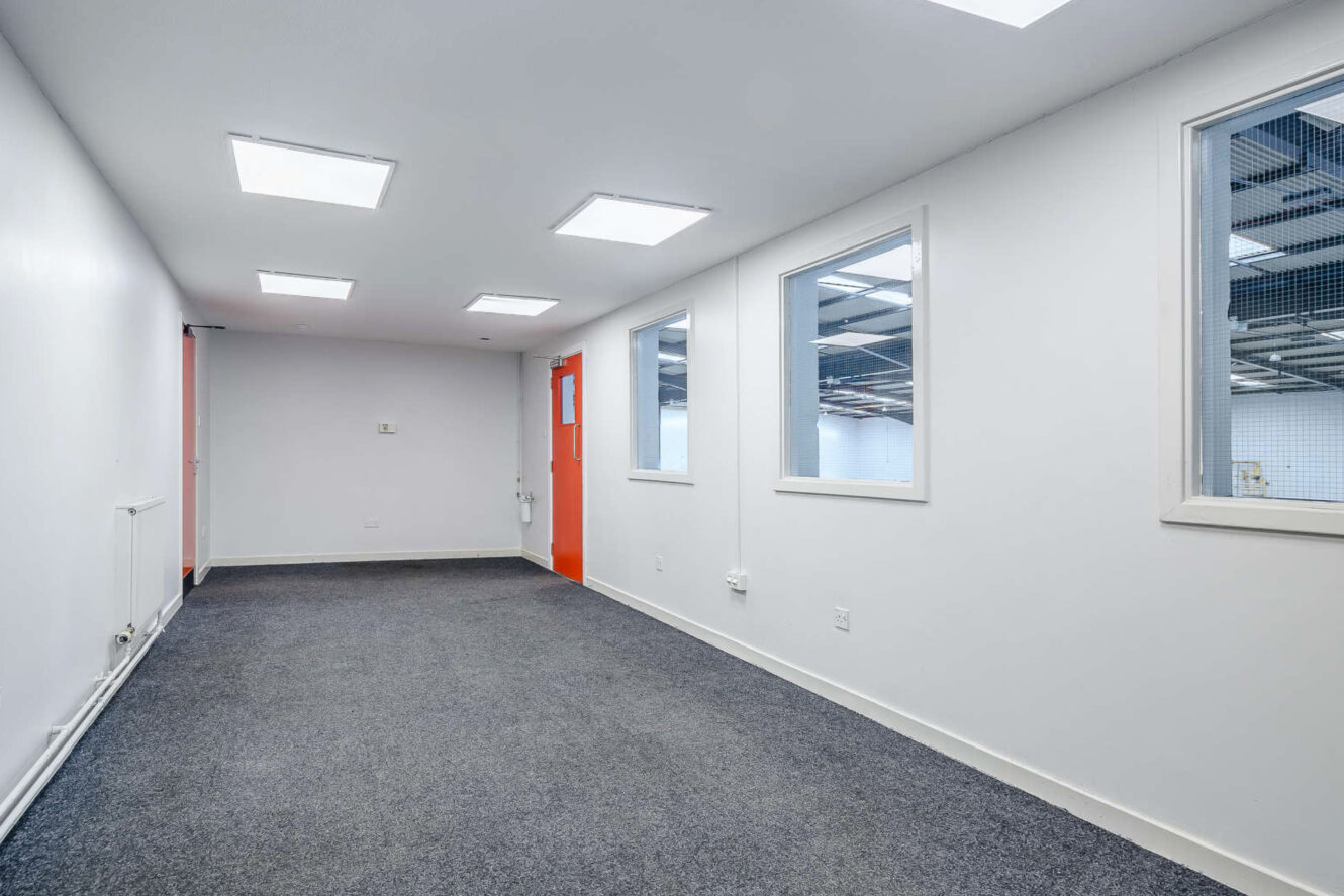 A clean, empty office space with carpeted flooring, white walls, large interior windows, and fluorescent ceiling lights. A red door leads to an adjacent industrial area.