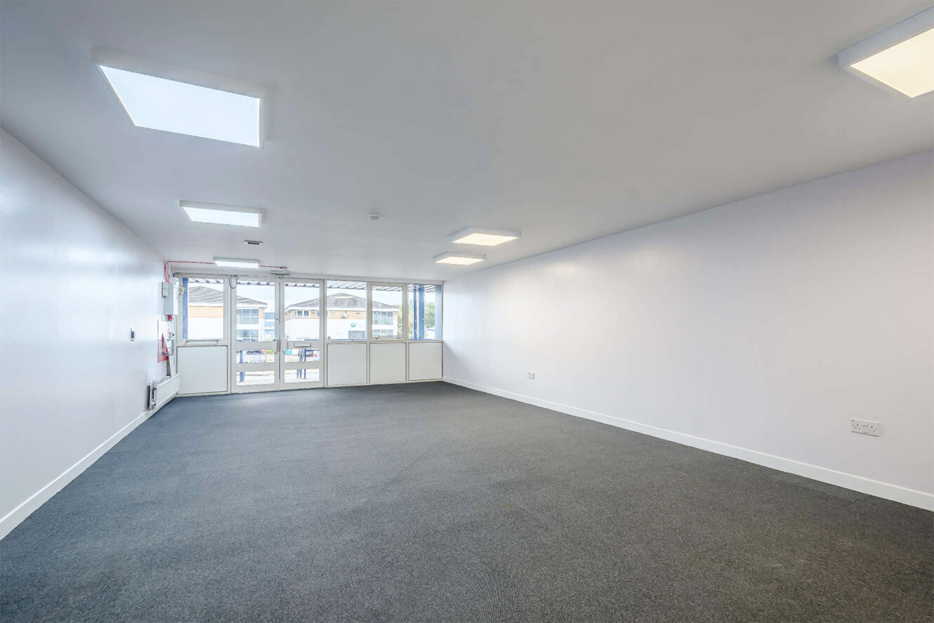 Empty office room with gray carpet, white walls, ceiling lights, skylights, and large windows facing outside.