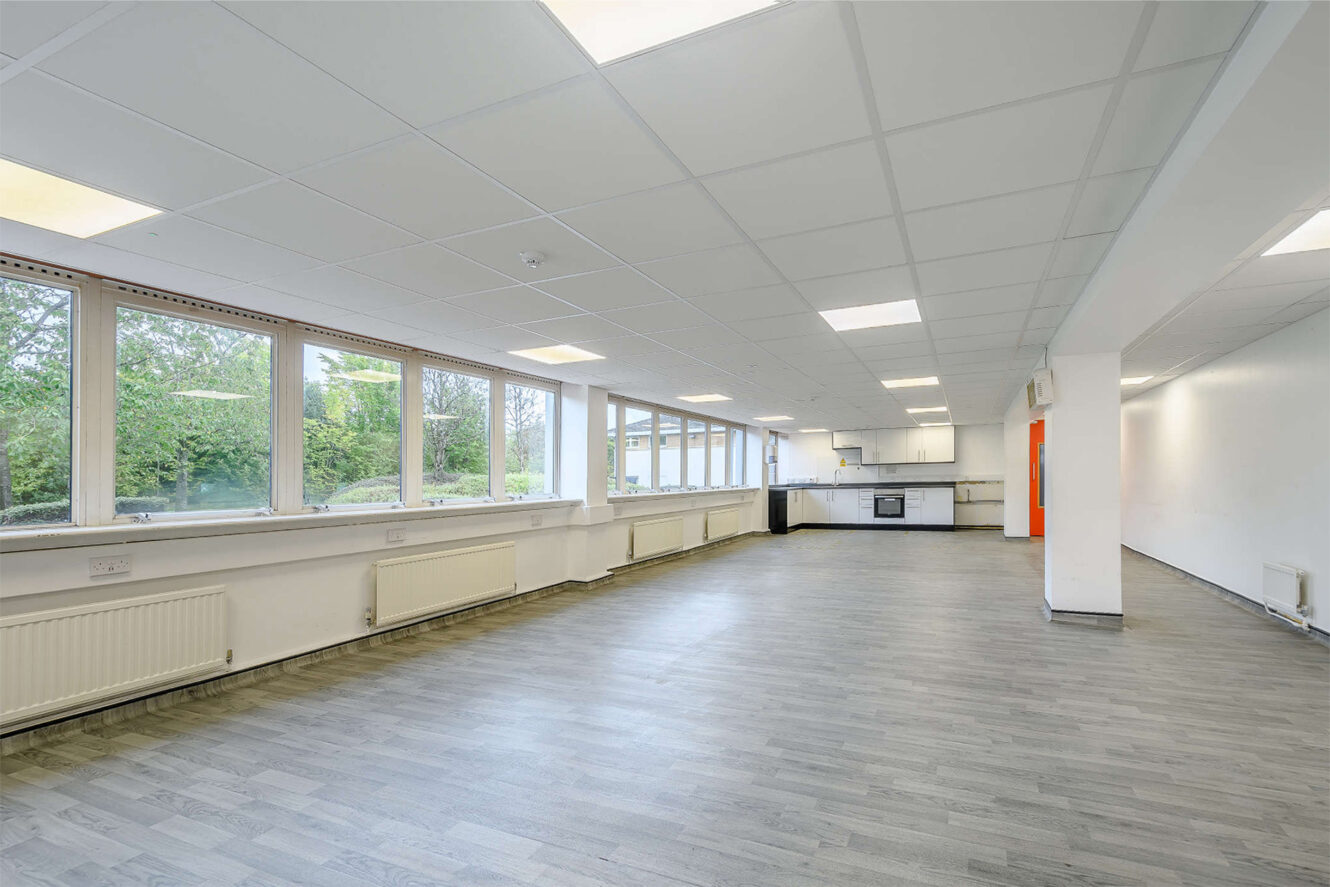 Spacious, empty room with large windows, grey flooring, white walls, and a kitchen area at the far end. Ceiling lights are on, and trees are visible outside.
