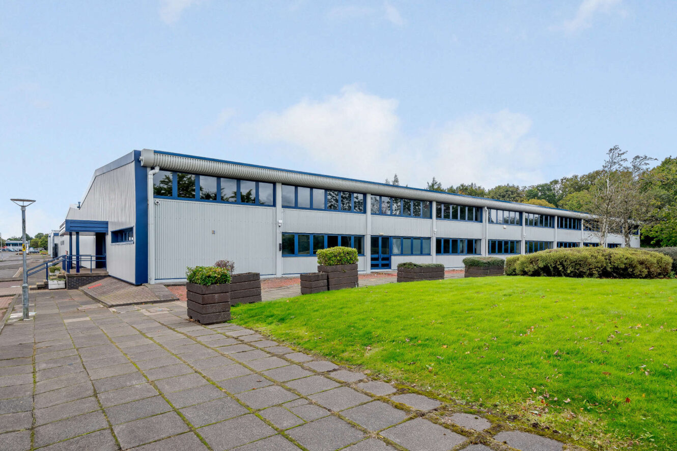 A modern, two-story industrial or office building with blue accents, large windows, and a landscaped lawn with shrubs and paving in the foreground.